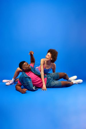 Foto de Mujer americana africana joven positiva con el maquillaje audaz en el vestido posando cerca de mejor amigo con estilo en chaleco de mezclilla y camiseta sobre fondo azul, besties de moda irradiando confianza - Imagen libre de derechos