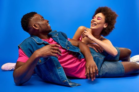 Téléchargez les photos : Souriant jeune femme afro-américaine avec un maquillage lumineux couché près du meilleur ami élégant en t-shirt et gilet en denim sur fond bleu, besties à la mode rayonnant de confiance - en image libre de droit