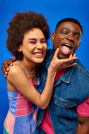 Porträt einer lächelnden jungen afrikanisch-amerikanischen Frau mit kühnem Make-up, die die trendige beste Freundin berührt, die die Zunge herausstreckt und isoliert auf blauen, modischen Bestien steht, die Zuversicht ausstrahlen 