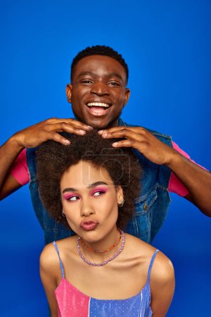 Smiling young african american man with modern hairstyle and summer outfit touching hair of best friend with bold makeup isolated on blue, fashionable besties radiating confidence, friendship
