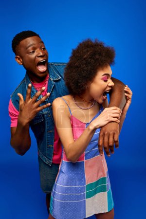 Photo for Young and trendy african american woman in stylish sundress biting arm of scared best friend in denim vest and standing isolated on blue, fashionable besties radiating confidence, friendship - Royalty Free Image