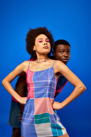 Confident young african american woman with bold makeup and sundress holding hands on hips while covering scared best friend isolated on blue, friends showcasing individual style, friendship