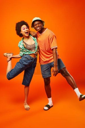Full length of cheerful young african american woman in trendy summer outfit hugging best friend in panama hat and looking at camera on orange background, friends showcasing individual style