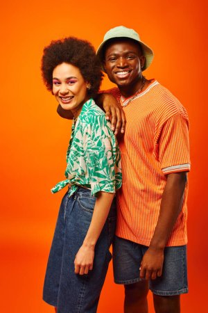 Smiling young african american man in panama hat and summer outfit looking at camera near best friend with bold makeup and standing isolated on orange, friends showcasing individual style