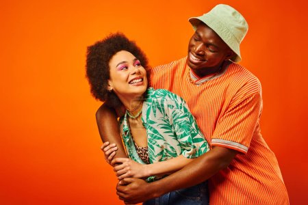 Pleased young african american man in panama hat embracing trendy best friend with bold makeup and looking at each other isolated on orange, friends showcasing individual style