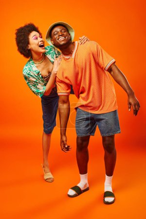Longitud completa de la joven afroamericana emocionada en traje de verano abrazando alegre mejor amigo en sombrero de panama mientras está de pie sobre fondo naranja, amigos mostrando el estilo individual