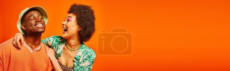 Excited young african american woman with bold makeup hugging cheerful best friend in summer outfit and panama hat isolated on orange, friends showcasing individual style, banner 