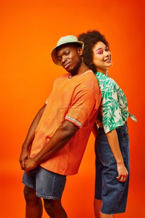 Junger afrikanisch-amerikanischer Mann im Sommer-Outfit und Panamahut blickt in die Kamera und steht Rücken an Rücken mit bester Freundin mit fettem Make-up und posiert isoliert auf orange, Freunde in trendigen Klamotten