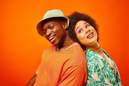 Fröhlicher junger afrikanisch-amerikanischer Mann mit Panamahut, der in die Kamera blickt, während er Rücken an Rücken mit seiner besten Freundin im Sommeroutfit zusammen isoliert auf orangefarbener Kleidung steht, Freunde in trendiger Kleidung