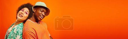 Junger und zufriedener afrikanisch-amerikanischer Mann in Panamahut und Sommerkostüm Rücken an Rücken mit bester Freundin mit fettem Make-up isoliert auf orange, Banner, Freunde in trendiger Kleidung, Freundschaft
