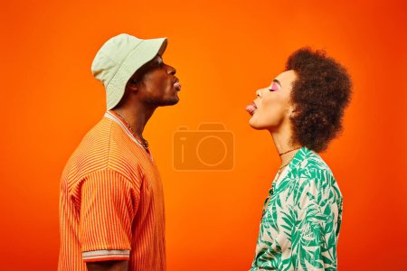 Photo for Side view of young and trendy african american best friends in summer outfits sticking out tongues while standing together isolated on orange, friends in trendy clothes, friendship concept - Royalty Free Image