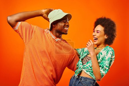Cheerful young african american man in panama hat hugging best friend with bold makeup and summer outfit and having fun together isolated on orange, friends in trendy clothes, friendship