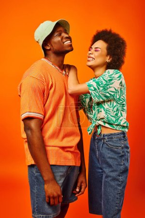 Positive junge afrikanisch-amerikanische Frau mit kühnem Make-up und sommerlichem Outfit umarmt stilvolle beste Freundin mit Panamahut, während sie isoliert auf orange steht, Freunde in trendigen Klamotten