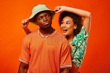 Smiling young african american woman with bold makeup in summer clothes touching panama hat of serious best friend and standing isolated on orange, friends in trendy clothes, friendship