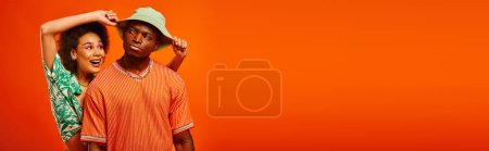 Positive junge afrikanisch-amerikanische Frau mit kühnem Make-up und sommerlichem Outfit, das Panamahut einer ernsthaften besten Freundin berührt, isoliert auf orangefarbenem Banner, modebewusste Freunde