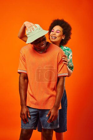 Positive junge afrikanisch-amerikanische Frau mit kühnem Make-up, die trendige beste Freundin mit Panamahut berührt, während sie isoliert auf orangefarbenen, modebewussten Freundinnen posiert, Freundschaft