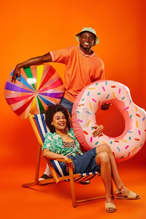 Full length of cheerful african american man in summer outfit holding pool ring and ball while standing near best friend on deck chair on orange background, fashion-forward friends, friendship