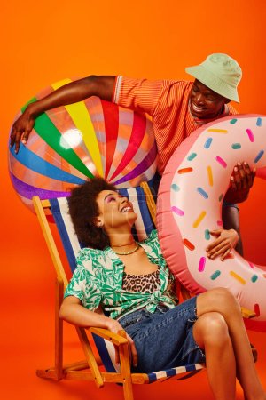 Positive young african american best friends in stylish summer outfit holding pool ball and ring near deck chair on orange background, fashion-forward friends, friendship concept 