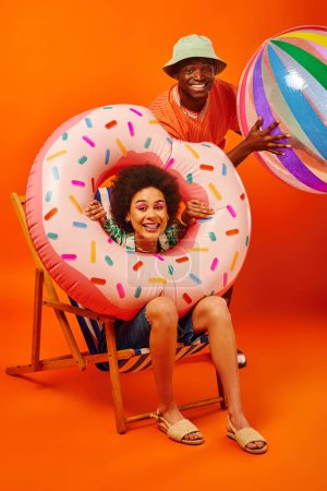 Positivo jóvenes afroamericanos mejores amigos en trajes de verano celebración de anillo de piscina y pelota mientras está sentado en la silla de cubierta y mirando a la cámara en el fondo naranja, amigos de moda hacia adelante