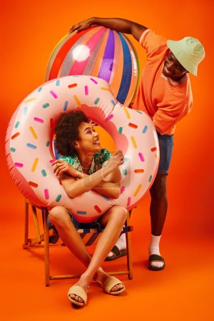 Joyeux jeune femme afro-américaine en tenue d'été tenant anneau de piscine tout en étant assis sur une chaise longue et en regardant meilleur ami avec balle sur fond orange, amis avant-gardistes de la mode