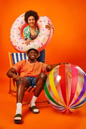 Full length of positive african american woman holding swim ring while standing near best friend in panama hat and summer outfit sitting on deck chair on orange background, fashion-forward friends