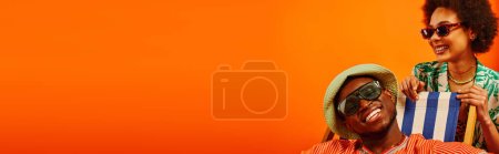 Foto de Sonriente y elegante mujer afroamericana joven en gafas de sol y traje de verano de pie cerca de mejor amigo en sombrero de panama sentado en la silla de cubierta aislado en naranja, amigos de moda hacia adelante, pancarta - Imagen libre de derechos