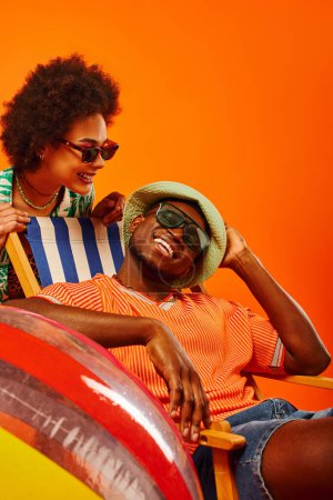 Pleased young african american woman in sunglasses and stylish summer outfit standing near best friend in panama hat relaxing on deck chair isolated on orange, friends in trendy casual attire