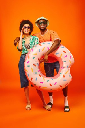Full length of smiling young african american man in sunglasses and panama hat hugging stylish best friend and holding swim ring on orange background, friends in trendy casual attire