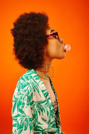 Téléchargez les photos : Vue latérale de jeune femme afro-américaine à la mode portant des lunettes de soleil en tenue d'été tout en soufflant de la gomme à bulles et se tenant isolée sur le rouge, femme avec un sens du style, confiance - en image libre de droit
