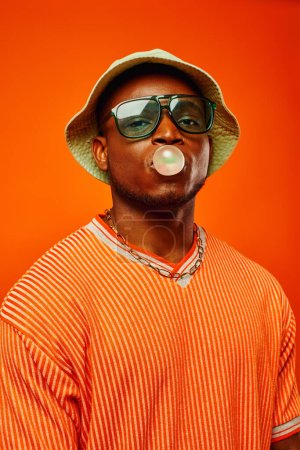 Retrato de hombre afroamericano joven de moda en gafas de sol y sombrero de panama que sopla goma de mascar y mirando a la cámara mientras está de pie aislado en rojo, hombre con sentido del estilo, confianza