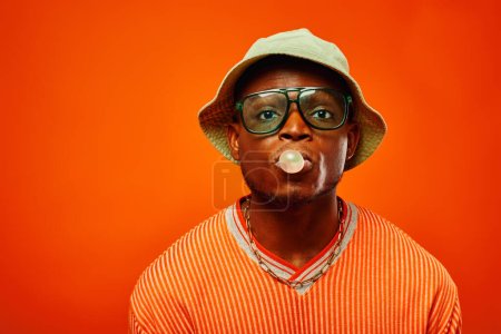 Photo for Portrait of young and stylish african american man in summer outfit wearing sunglasses and panama hat blowing bubble gum and looking at camera isolated on red, man with sense of style - Royalty Free Image