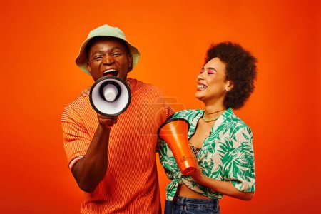 Cheerful young african american woman with bold makeup and summer outfit looking at best friend in panama hat screaming at loudspeaker isolated on red, friends in fashionable outfits
