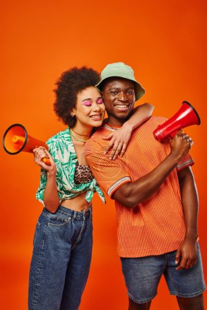 Smiling young african american woman with bold makeup and summer outfit hugging best friend in panama hat and holding loudspeaker while standing isolated on red, friends in fashionable outfits
