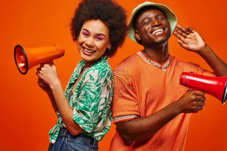 Positive young african american best friends in summer clothes looking at camera while holding loudspeakers and standing isolated on red, friends in fashionable outfits, friendship