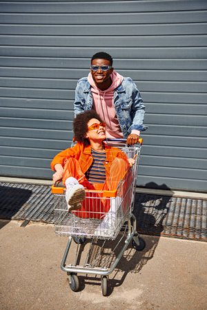 Smiling young african american man in denim jacket and sunglasses standing near best friend in shopping cart and building on urban street, friends hanging out together, friendship