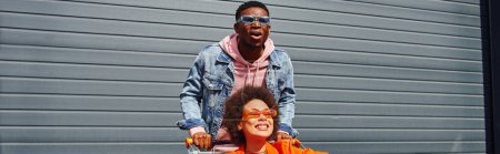 Excited young african american man in sunglasses and denim jacket standing near positive best friend sitting in shopping cart and building at background, friends hanging out together, banner 