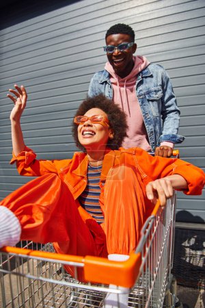 Cheerful young african american woman in bright outfit and sunglasses sitting in shopping cart and having fun with best friend near building on urban street, friends hanging out together