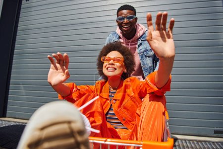 Positive junge afrikanisch-amerikanische beste Freunde in Sonnenbrille und hellem, stylischem Outfit haben Spaß mit Einkaufswagen in der Nähe von Gebäuden im Hintergrund auf der urbanen Straße, Freunde hängen zusammen