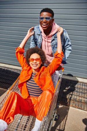 Positive and fashionable african american best friends in sunglasses and bright clothes looking at camera and having fun with shopping cart on urban street, friends hanging out together