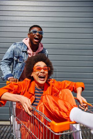 Aufgeregt junge und trendige afrikanisch-amerikanische beste Freunde in Sonnenbrille und heller Kleidung haben Spaß mit Einkaufswagen in der Nähe Gebäude im Hintergrund auf der städtischen Straße, Freunde hängen zusammen