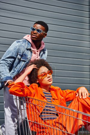 Mujer afroamericana joven de moda en gafas de sol y traje brillante sentado en el carrito de la compra cerca de mejor amigo confiado en chaqueta de mezclilla y la construcción de fondo, amigos pasando el rato juntos