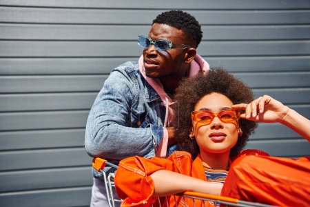 Confident and trendy young african american best friends in sunglasses and bright clothes posing with shopping cart near building at background on urban street, friends with stylish vibe