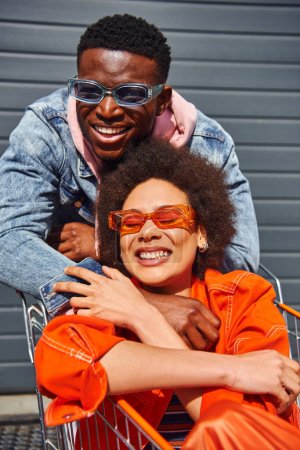 Portrait of cheerful young african american man in sunglasses and denim jacket looking at camera while hugging best friend in shopping cart near building on urban street, friends with stylish vibe