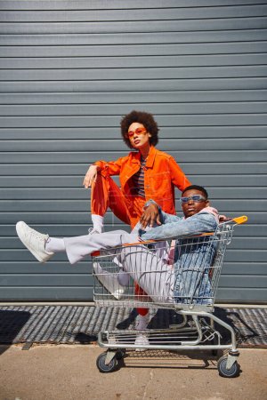 Full length of trendy young african american woman in sunglasses and bright outfit posing near best friend sitting in shopping cart and looking at camera on urban street, friends with stylish vibe
