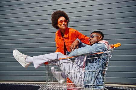 Stylish young african american woman in sunglasses and bright outfit looking away near best friend in denim jacket sitting in shopping cart and building outdoors, friends with stylish vibe
