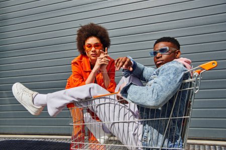 Téléchargez les photos : Confiants et branchés jeunes afro-américains meilleurs amis dans des lunettes de soleil et des tenues lumineuses posant avec panier près du bâtiment dans la rue urbaine, amis avec le concept d'ambiance élégant - en image libre de droit