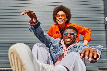 Téléchargez les photos : Femme afro-américaine positive dans des lunettes de soleil et des vêtements lumineux s'amuser avec le meilleur ami élégant dans la veste en denim assis dans le panier dans la rue urbaine, amis avec ambiance élégante - en image libre de droit