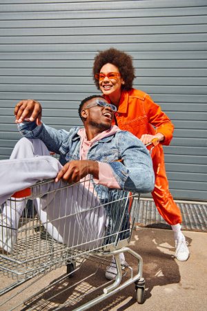 Fröhliche junge afrikanisch-amerikanische Frau mit Sonnenbrille und lebendigem Outfit, die Spaß mit ihrer stilvollen besten Freundin hat, die im Einkaufswagen sitzt und Zeit auf urbanen Straßen verbringt, Freundinnen mit stilvollem Ambiente