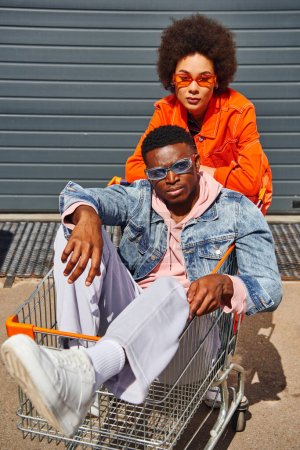 Fashionable african american woman in sunglasses and bright clothes looking at camera while standing near confident best friend sitting in shopping cart on urban street, stylish friends in city
