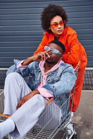 Modern young african american woman in sunglasses and bright outfit looking away while standing near best friend sitting in shopping cart and building on urban street, stylish friends in city